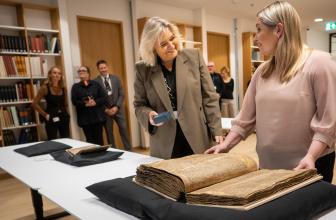 Manuscripts rest on a table, on black pillows. A woman in a pink sweater stands at the largest manuscript, another woman in a blazer stands next to her and smiles at her. A few people look on from the background, standing in front of bookshelves..
