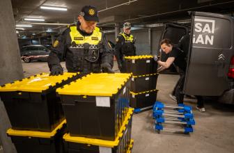 Grey, concrete car park. A man pushes a stack of yellow and black boxes from the back of a black van. Two stacks of boxes are already on the floor and two policemen watch the proceedings.