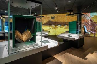 Overview inside exhibition space. On top of white tables are glass cases. Within them are green boxes that frame old manuscripts. The far wall is filled from floor to ceiling with drawn images of landscapes, foliage and a raven in flight.