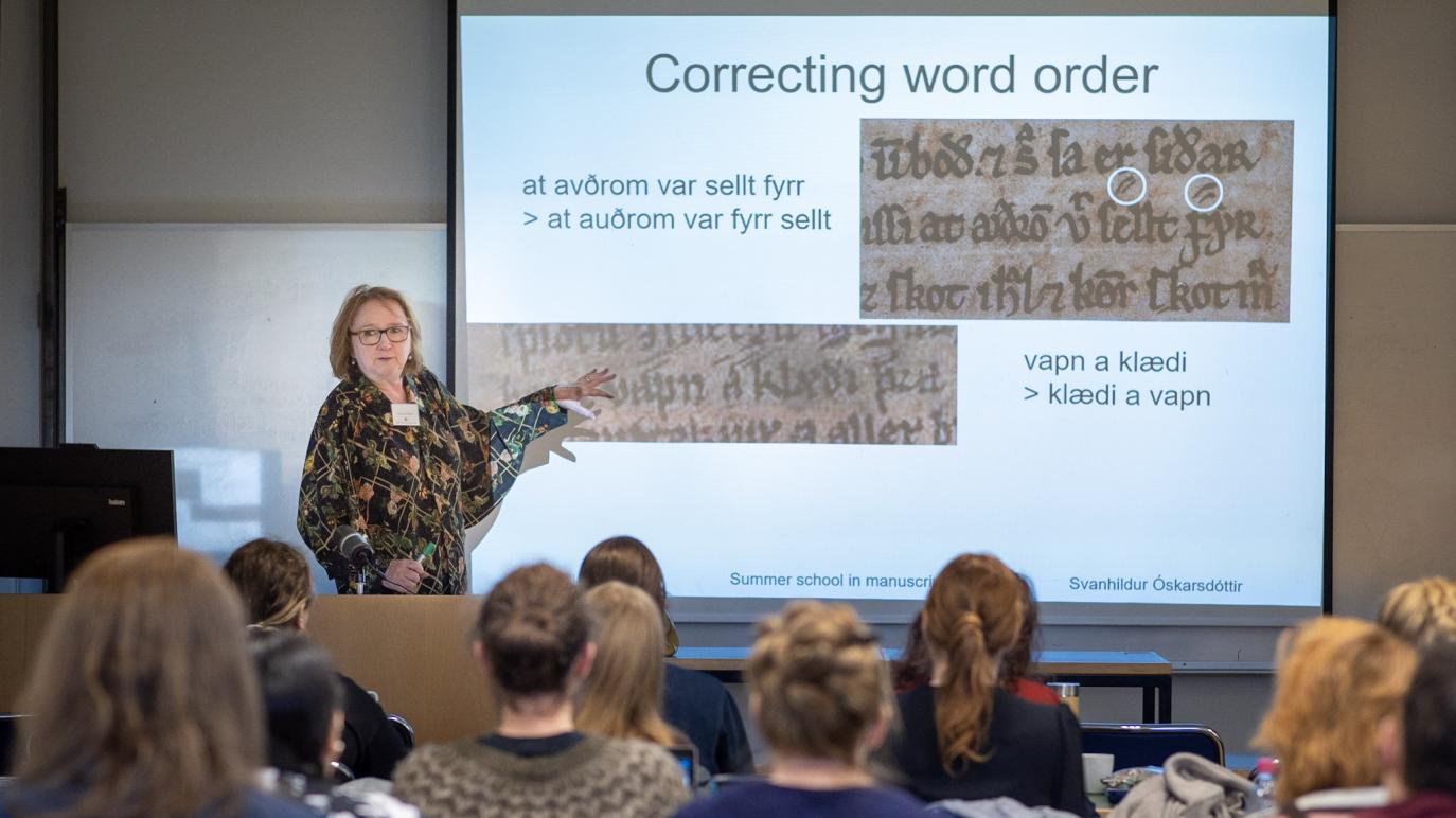 Kona með gleraugu í skrautlegri, svartri og grænni skyrtu stendur við púlt og bendir á mynd af handritasíðu á stórum skjá. Fyrir framan hana situr hópur nemenda og skýr baki í myndavélina.