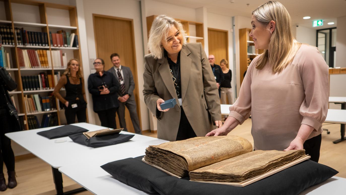 Manuscripts rest on a table, on black pillows. A woman in a pink sweater stands at the largest manuscript, another woman in a blazer stands next to her and smiles at her. A few people look on from the background, standing in front of bookshelves..