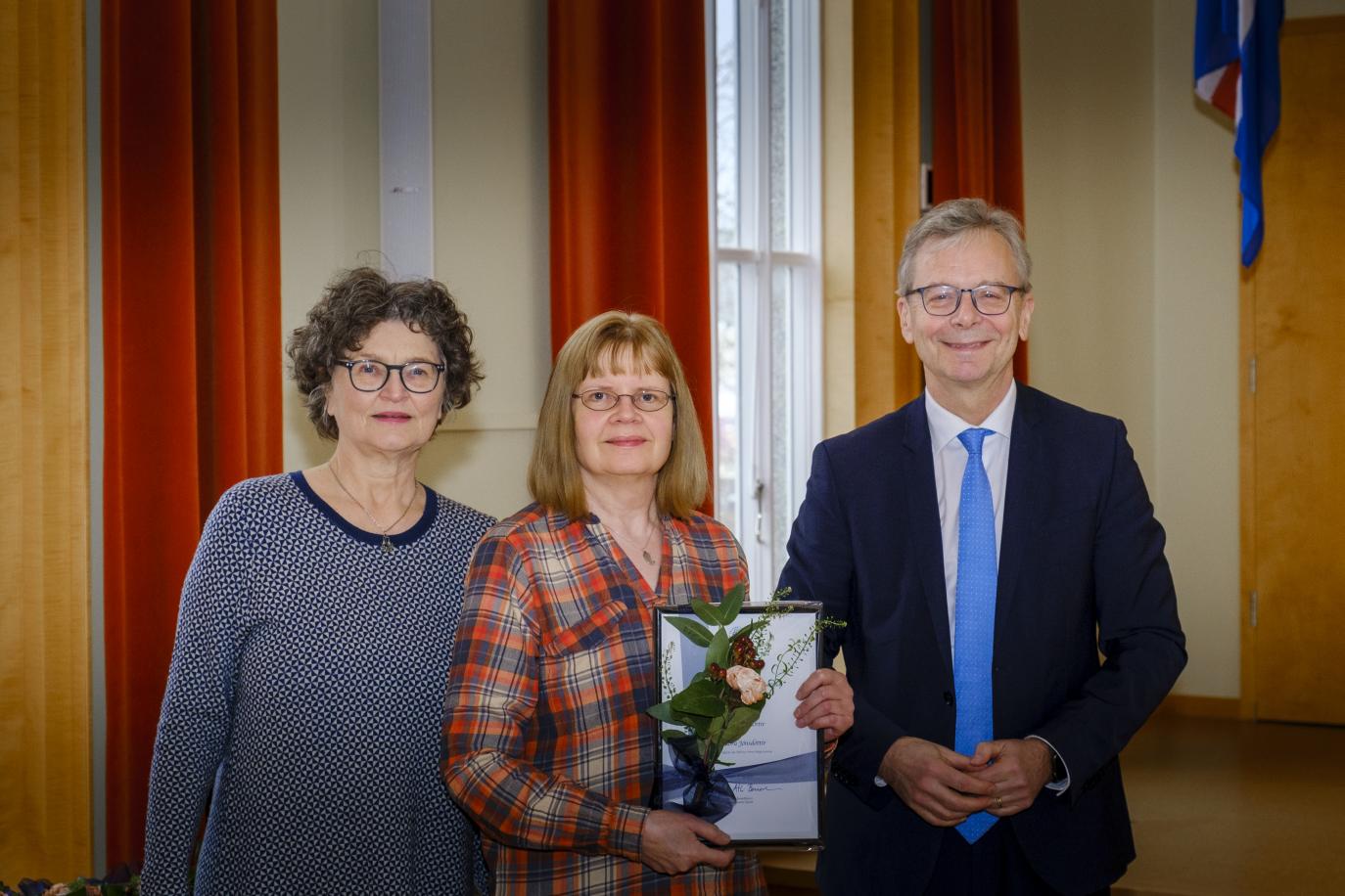 Tvær konur og karl. Kona í miðjunni heldur á viðurkenningarskjali. Rauðleit gluggatjöld í bakgrunni.