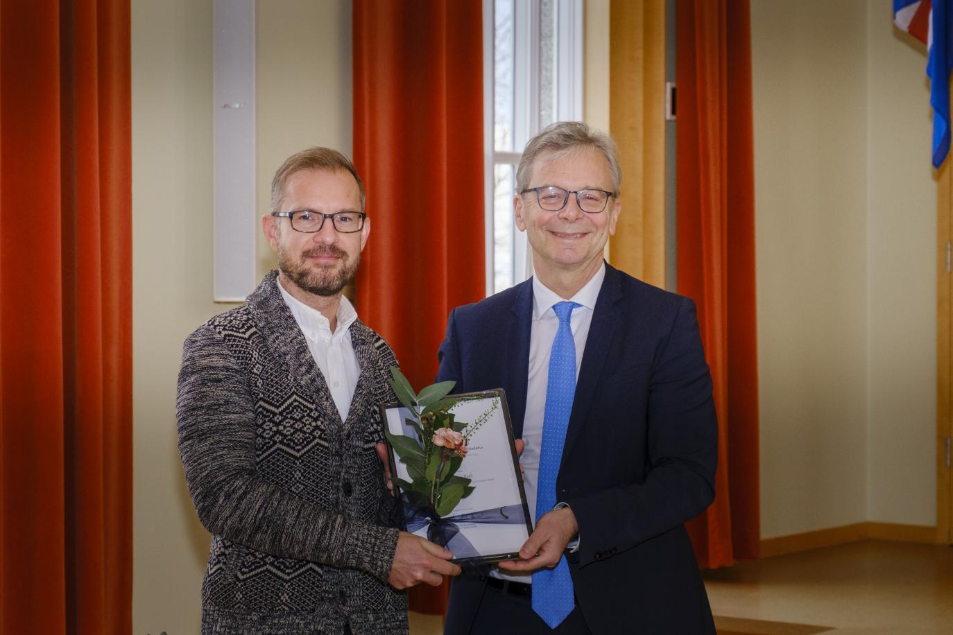 Tveir menn halda á viðurkenningarskjali. Rauðleit gluggatjöld í bakgrunni.