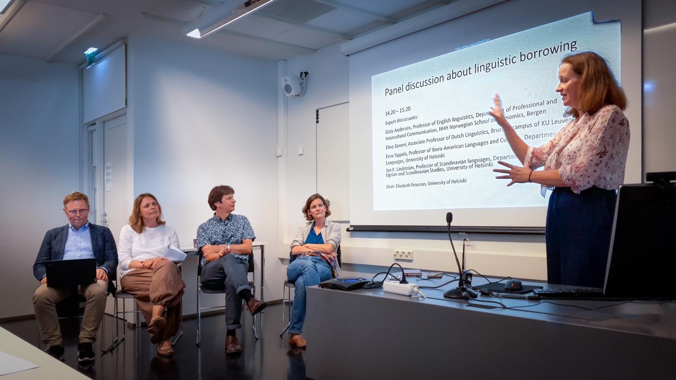 Kona stendur við skrifborð og fjórar manneskjur sitja í röð við hlið hennar. Á skjá stendur "Panel discussion about linguistic borrowing".