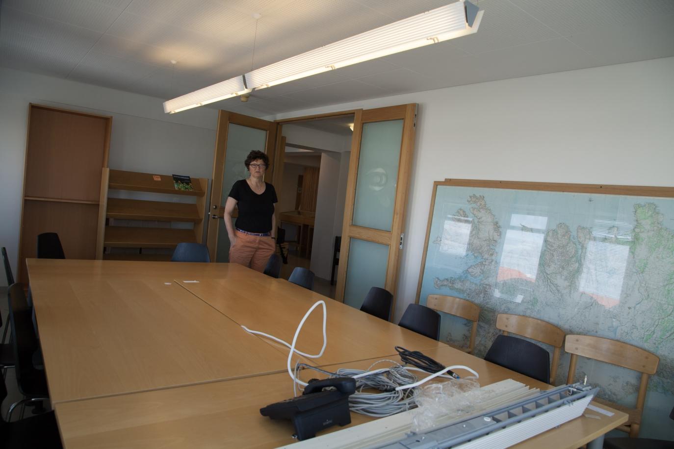 Kona stendur inn í fundarherbergi, á fundarborði í miðju herbergi eru rafmagns snúrur. bókahilla stendur í enda á herbergi tóm.