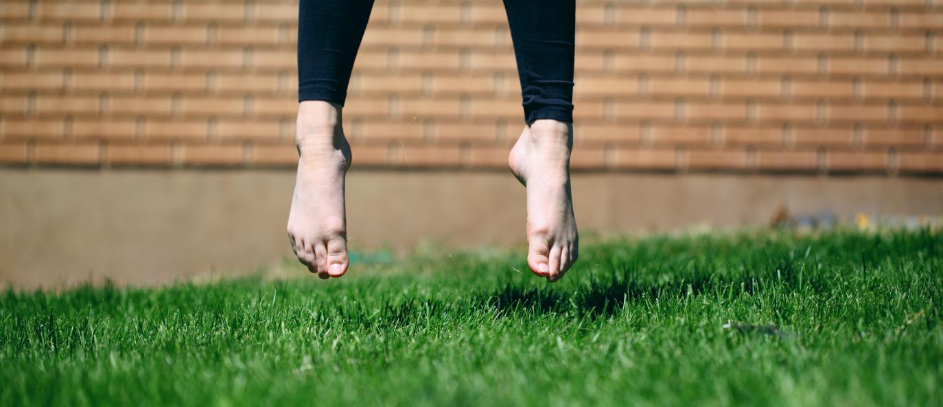 Fótleggir í miðju hoppi yfir grasfleti. Íklæddir leggings með bera fætur. Í bakgrunni er múrsteinsveggur, úr fókus.