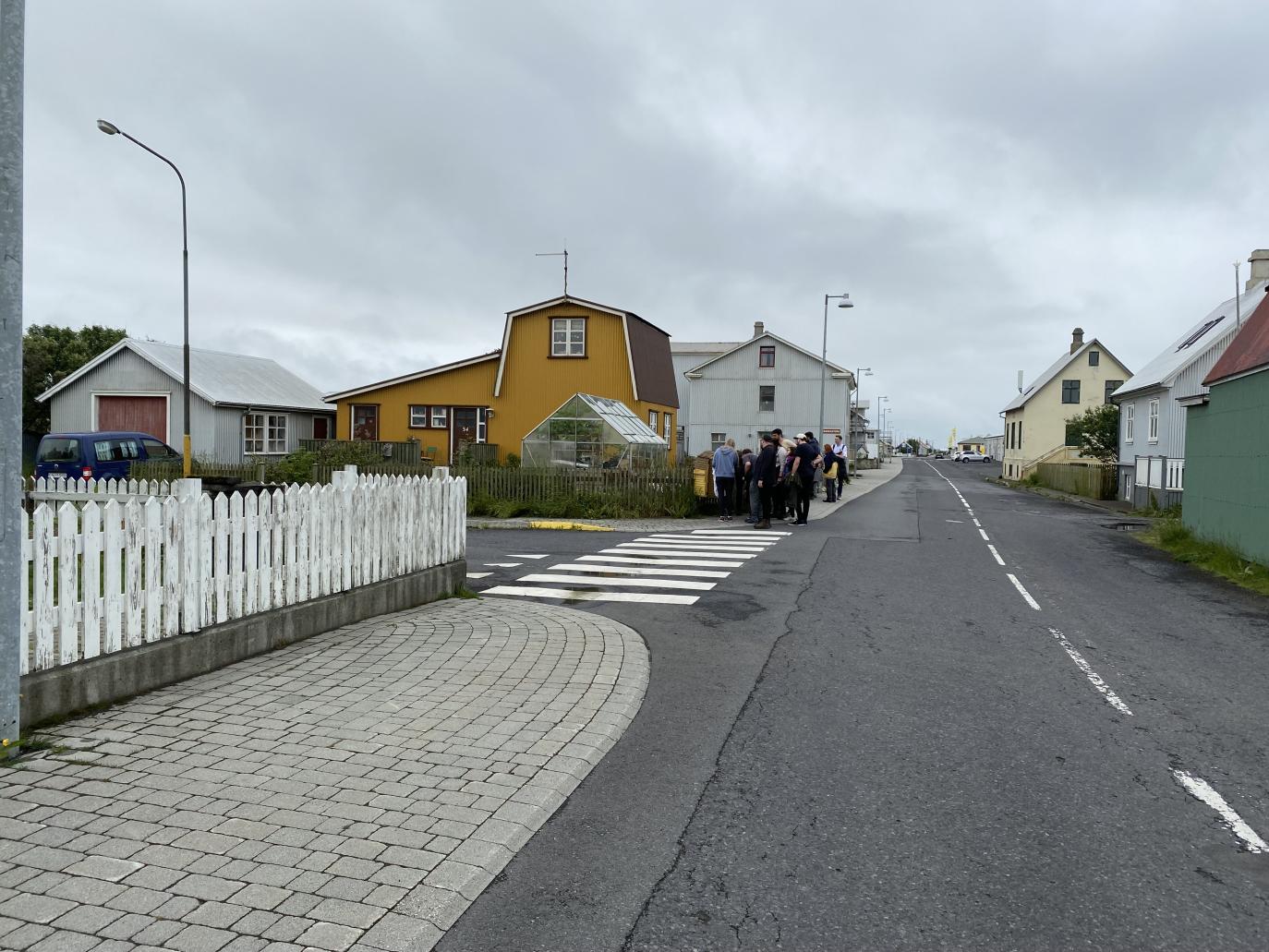 Horft eftir götu og hellulagðri gangstétt. Meðfram gangstéttinni er hvítt rimlagrindverk. Fjær sjást gömul hús sem eru hvít, gul og græn.