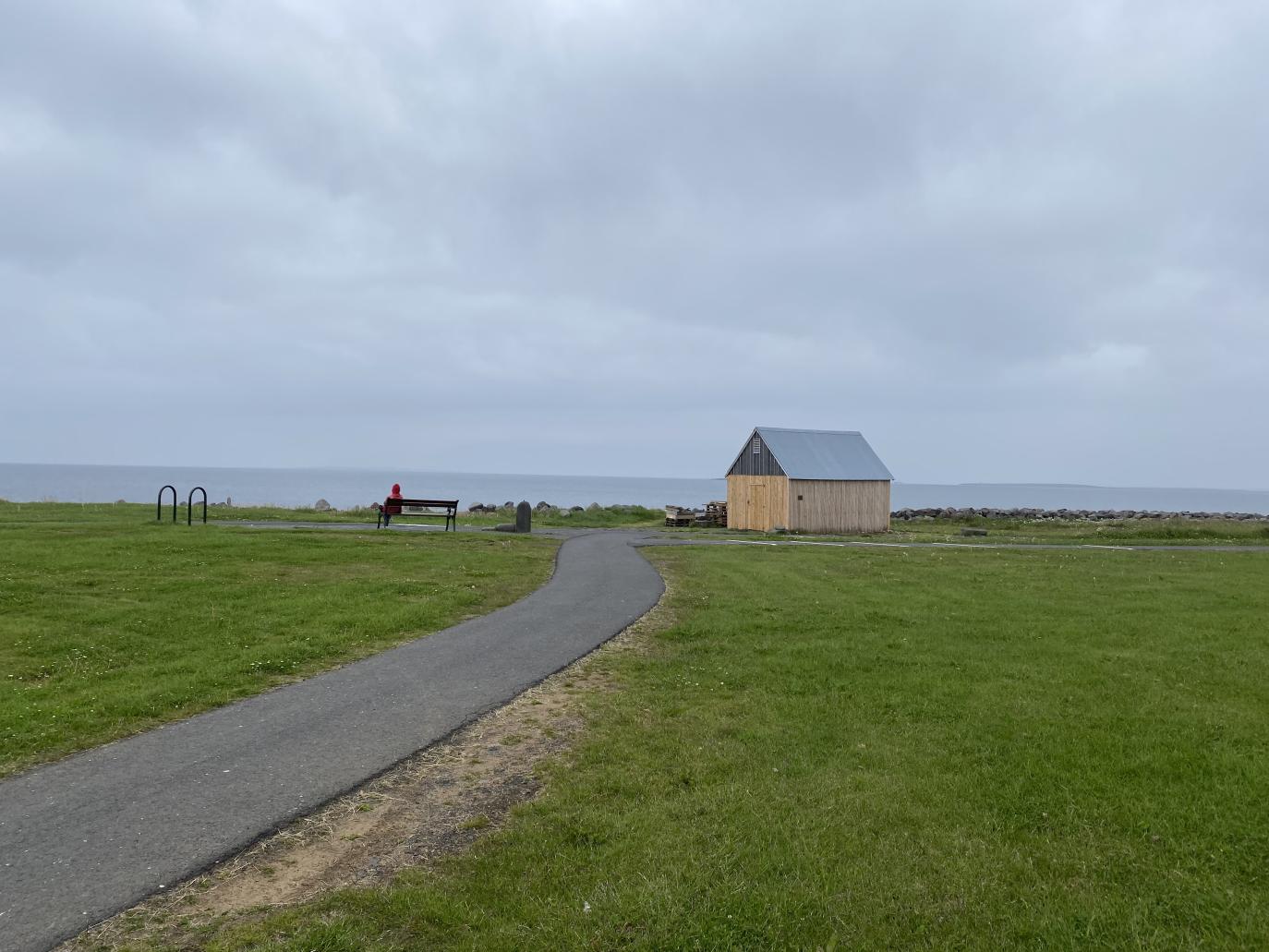 Göngustígur liggur að litlu gulu húsi með grátt þak sem stendur eitt með gras í kring. Fjær sést í sjóinn.