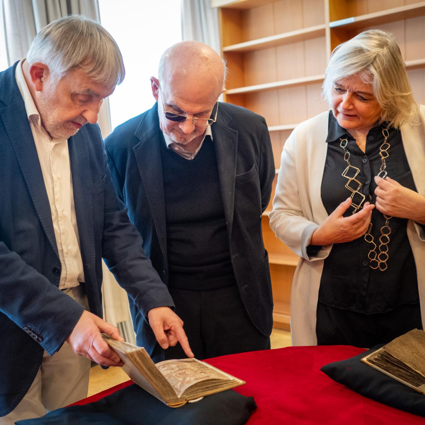 A grey-haired man in a dark blazer and a light-coloured shirt and trousers displays a manuscript resting on a black pillow. At his side is a bald man with glasses (one glass tinted) and a woman with shoulder-length hair, wearing a white coat over black clothes.