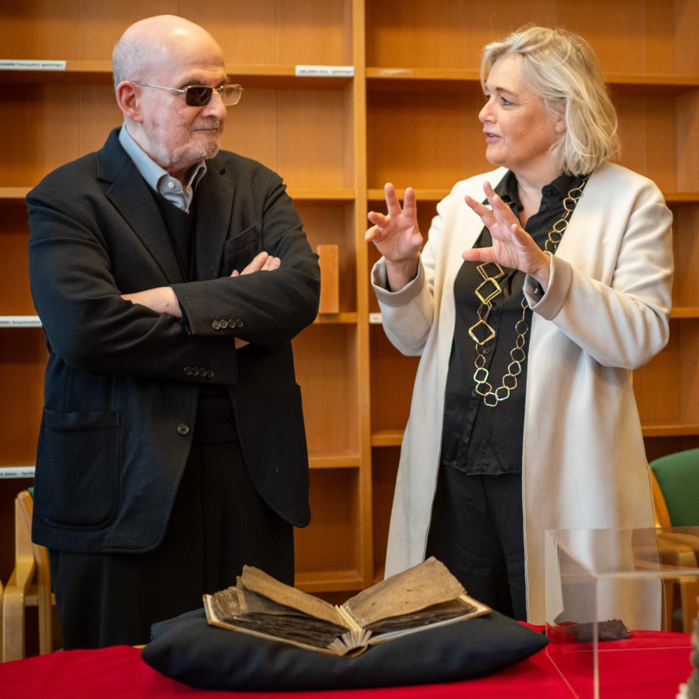 Bald man wearing a dark suit and glasses with one glass tinted and a woman with shoulder-length hair, wearing a white coat over a black dress are having a conversation. A medieval manuscript rests on a black pillow on the table in front of them.
