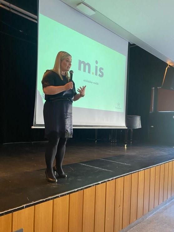 Woman in a black dress stands on a stage. Behind her, a green PowerPoint presentation reads "m.is.".