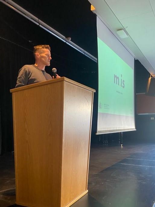 A man stands at a lectern. Beside him on a screen, a PowerPoint presentation reads "m.is".