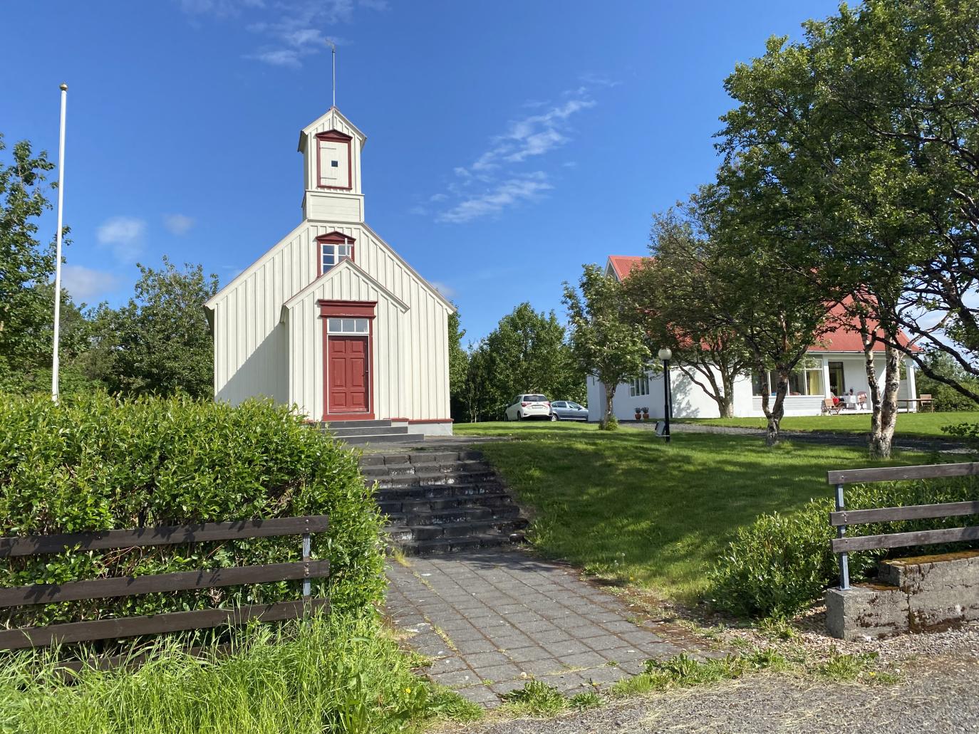 Hellulagður göngustígur liggur í gegnum garð að gamalli, hvítri kirkju með rauða hurð og gluggakarma.