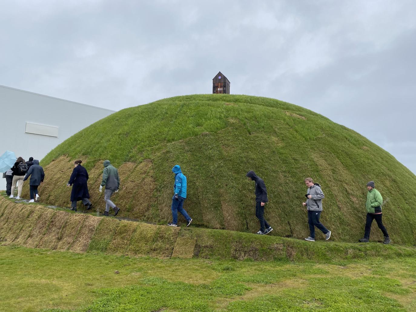 Grasi vaxinn, ávalur (manngerður) hóll með lítill byggingu á toppnum. Fólk gengur upp göngustíg sem fylgir útlínum hólsins.