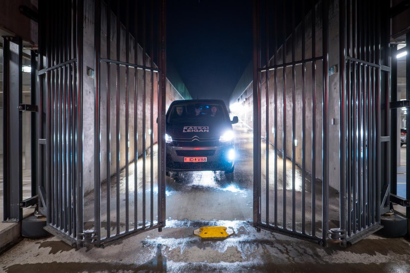 A tall gate folds open. Behind it is a black van, its lights illuminate the dark outside.