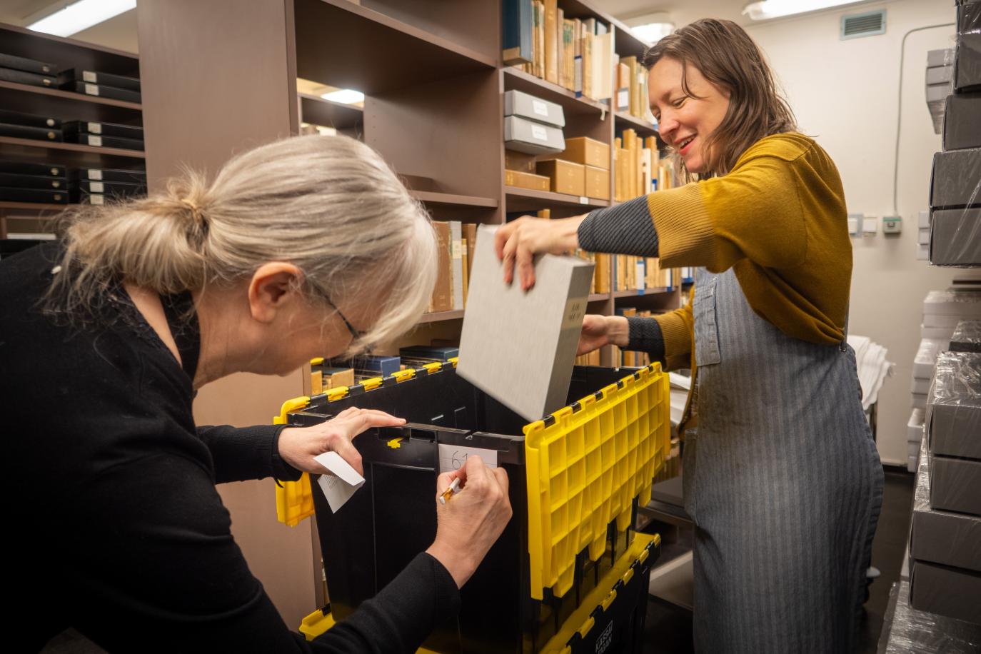 A woman in a yellow sweater and grey apron puts book into a yellow and black plastic box. Another woman in a black sweater write something on the outside of the box. Around them are shelves with boxes and books of various sizes.