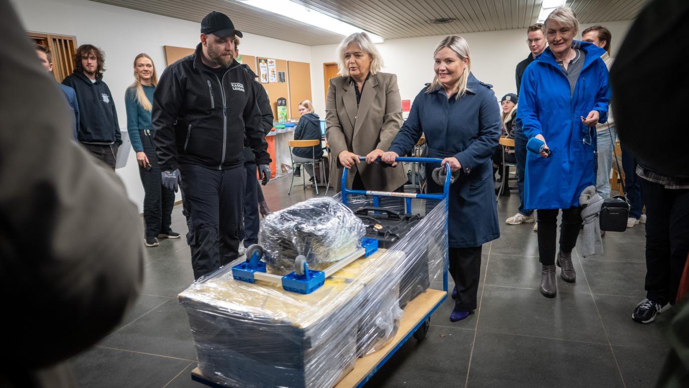 Two women push a cart, on it are boxes wrapped in plastic. A few people stand around and look on.