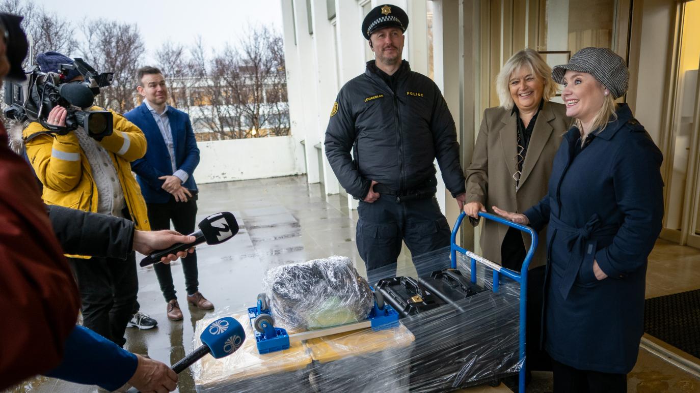 Two women push a cart with boxes wrapped in plastic and speak to reporters with cameras and microphones. Beside the women is a police officer.