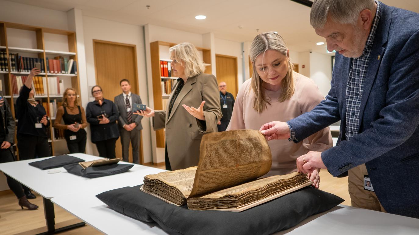 A man and a woman inspect a sizable, old manuscript that rests on a black pillow on top of a desk.. Behind them a woman talks and gesticulates with her hands. Further away, people stand in front of book cases and look on.