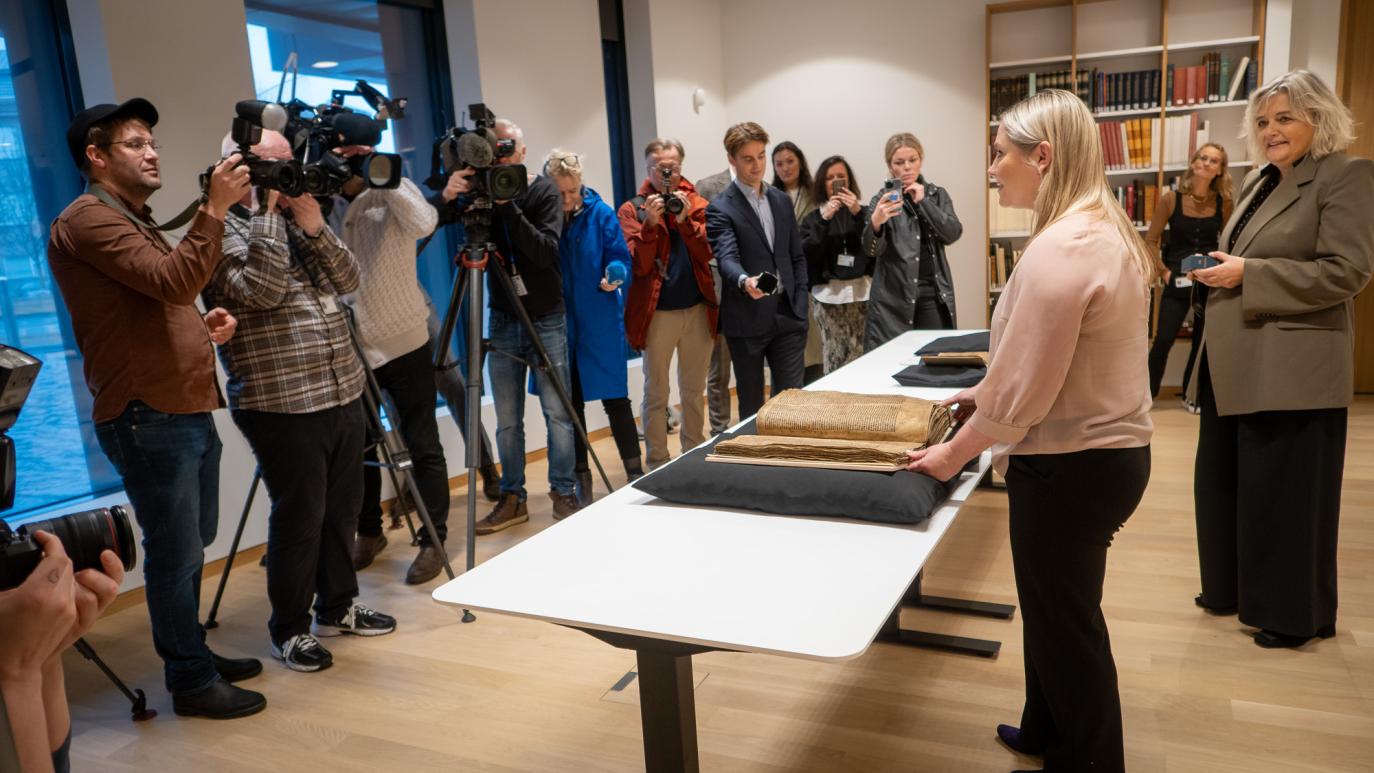 Two women stand in front of desks on which two manuscripts rest. In front of them are people from the media with cameras and microphones.