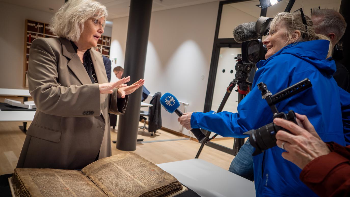 A womand stands in front of a manuscripts and speaks into a blue microphone held by a woman from the media. Beside the media woman is a camera and a cameraman.