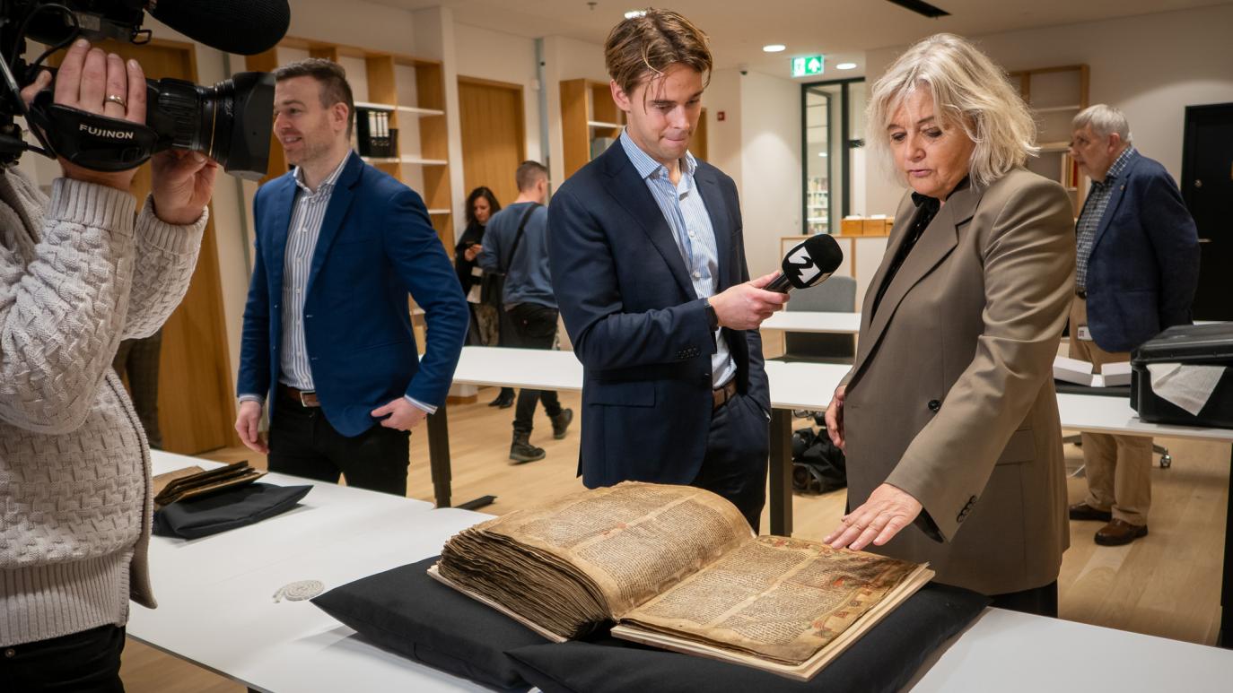 A woman points at a manuscript and speaks to a reporter who stands next to her. To the left, in the foreground, we see a cameraman pointing his camera at the pair.