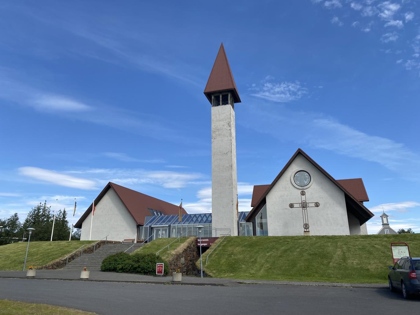 Hvítt hús með brúnu þaki, við það stendur hár, mjór turn. Fyrir framan húsið er stór kross.