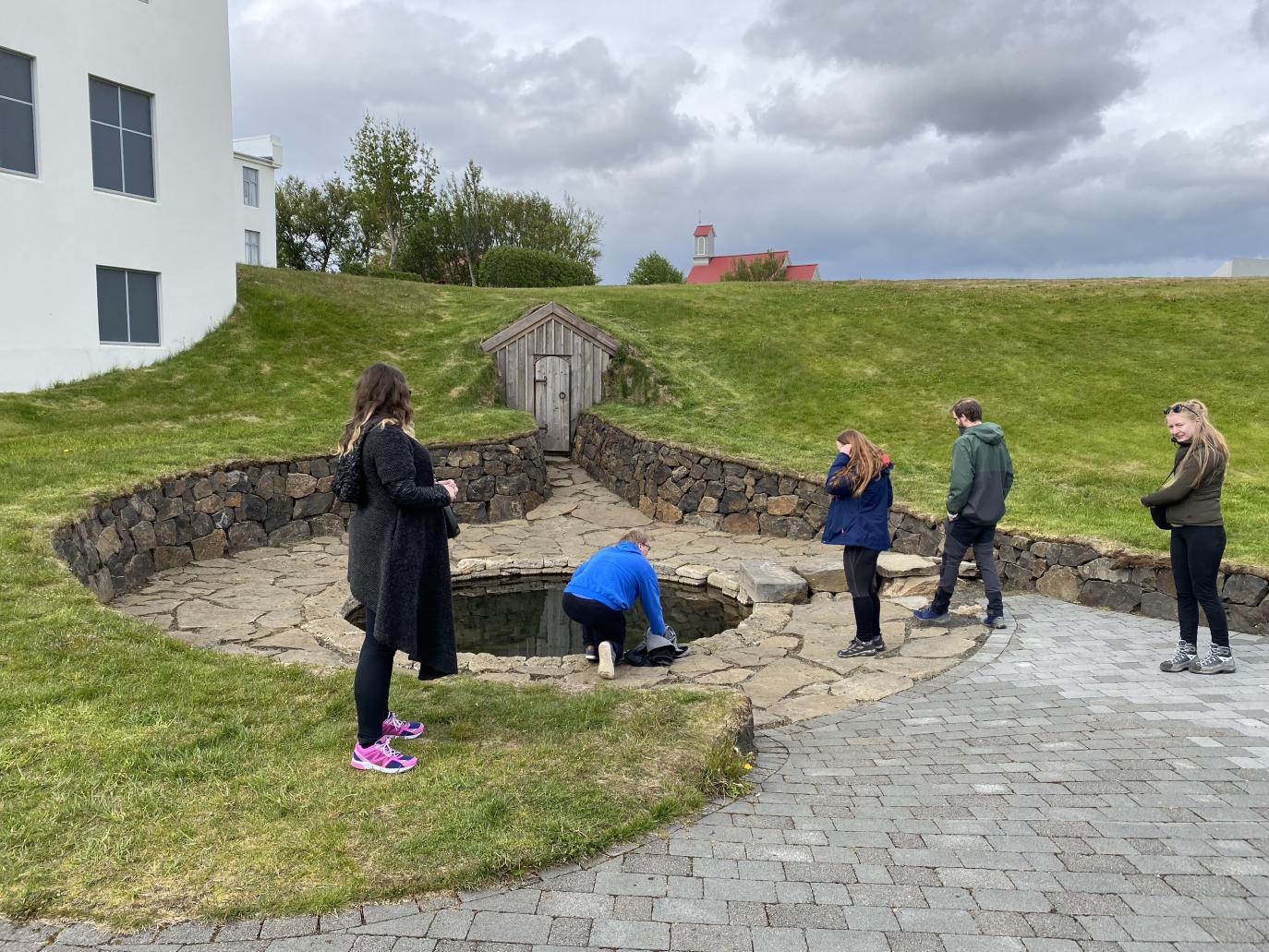 Hringlaga laug sem römmuð er inn með snyrtilegri hleðslu. Til hliðar, hvítt hús.