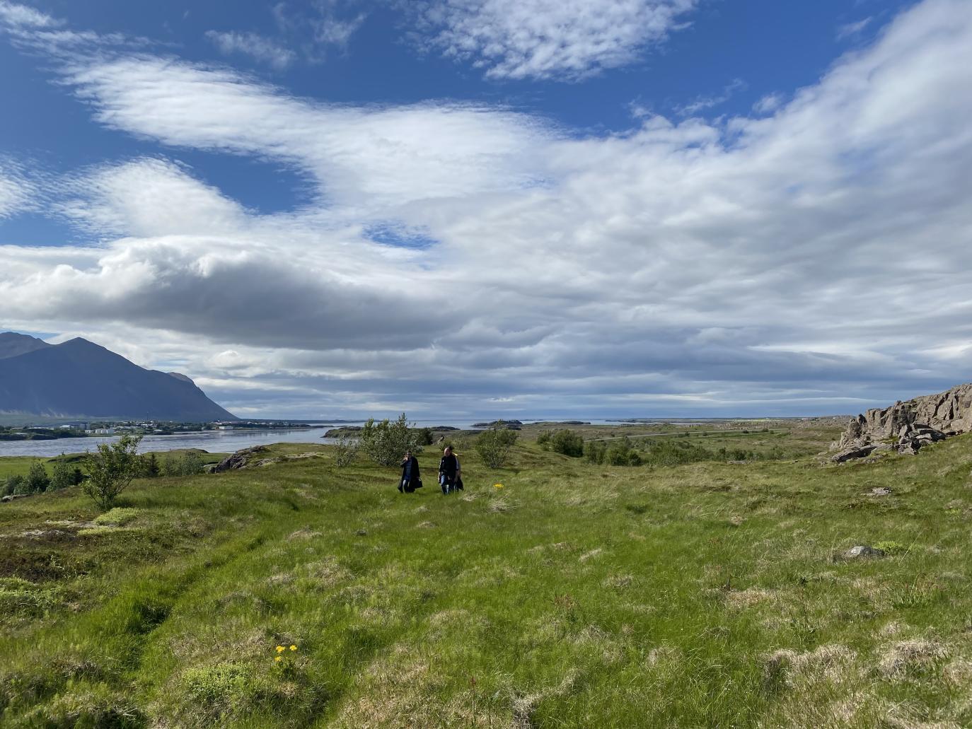 Þýft graslendi, ský á bláum himni og fjöll í fjarska. Fólk á gangi í átt að myndavél.