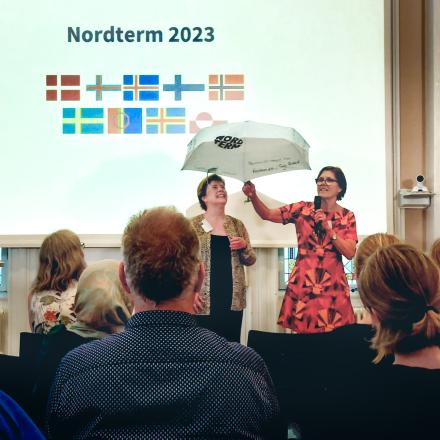 Tvær konur á sviði, önnur heldur á regnhlíf. Fyrir aftan þær á stórum skjá stendur "Nordterm 2023" og fyrir neðan það allir fánar Norðurlanda.