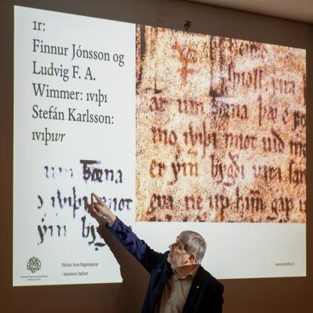 A grey-haired man in a dark blazer and white shirt points to old lettering on a PowerPoint slide. 