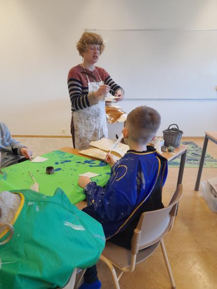 Kona með svuntu stendur og skrifar með fjöður. Börn í málningarsvuntum fylgjast með.